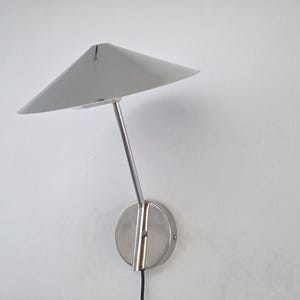 Könnte beinhalten: Eine moderne, silberfarbene Wandleuchte mit dreieckigem Schirm. Die Lampe hat ein schlankes, minimalistisches Design mit einem gebogenen Arm und einem kreisförmigen Sockel. An einer weißen Wand befestigt.