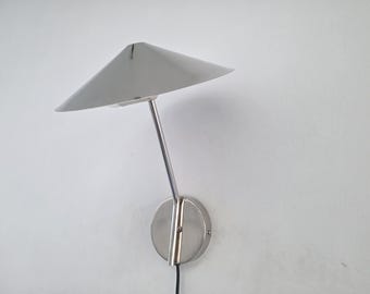 May include: A modern, silver-toned wall-mounted lamp with a triangular shade. The lamp features a sleek, minimalist design with a curved arm and a circular base. The shade is angled, and the lamp is mounted on a white wall.