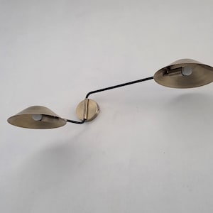 May include: A wall-mounted, two-light fixture with a brass finish. The fixture has two conical shades, a circular base, and a black arm. The light bulbs are visible inside the shades. The fixture is mounted on a white wall.
