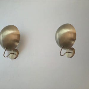 May include: Two brass wall sconces with a half-sphere shade and a curved arm. The sconces are mounted on a white wall. The sconces have a mid-century modern design.
