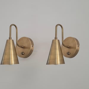 Op de afbeelding: Twee messing wandlampen met een conische kap en een gebogen arm. De lampen hebben een ronde basis en een warme, metallic afwerking. Het ontwerp is minimalistisch en modern, geschikt voor binnenverlichting.