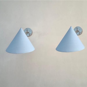 May include: Two light blue cone-shaped wall sconces with a circular decorative element. The sconces are mounted on a light beige wall. The sconces have a simple, modern design, and the light blue color provides a soft, ambient glow.