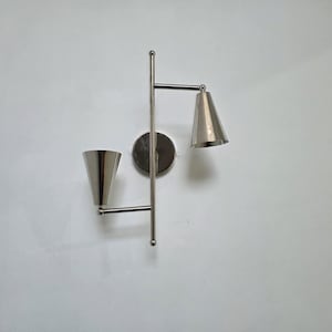 Op de afbeelding: Een moderne wandlamp met een geborstelde metalen afwerking. De lamp heeft twee conische kappen en een cirkelvormige achterplaat. De lamp is gemonteerd op een witte muur en biedt een strakke en eigentijdse uitstraling.