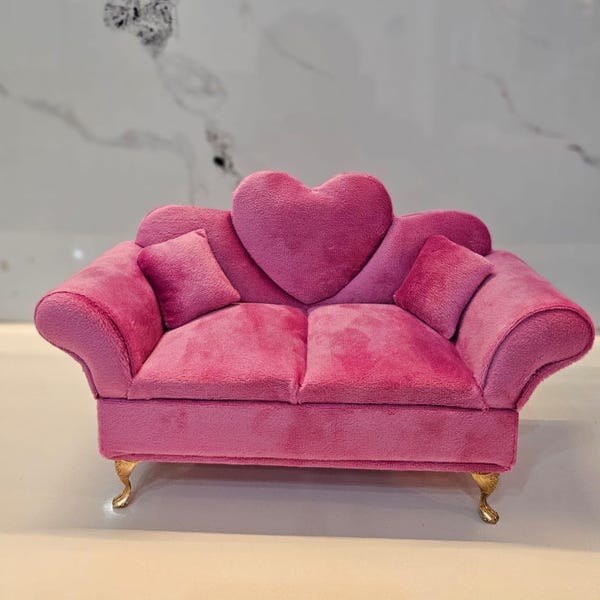 May include: A miniature sofa upholstered in bright pink plush fabric featuring a prominent heart-shaped backrest, two matching square throw pillows, and four ornate gold-toned cabriole legs. The piece has rolled arms and a two-cushion seat.