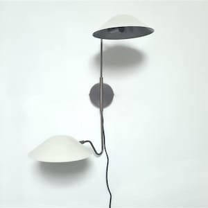May include: A contemporary wall light featuring two white, dish-shaped shades. The shades are attached to a dark metal frame with a circular base and a black power cable. The light is designed for indoor use and provides ambient illumination.