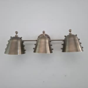 May include: A bronze-toned, three-light vanity fixture. Each bell-shaped shade is adorned with small, decorative rivets and topped with a small sphere. The fixture is mounted on a wall, showcasing a vintage or industrial design.