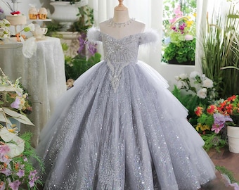 May include: A silver-gray off-the-shoulder dress with a layered skirt and sparkling embellishments. The bodice is intricately detailed with beadwork and feather accents. A delicate necklace adorns the neckline. The dress is displayed on a mannequin.