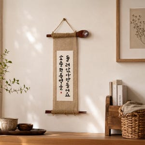 May include: A vertical wall hanging featuring a rectangular piece of tan burlap fabric suspended by a dark wooden spoon at the top and a pair of chopsticks at the bottom. A white paper strip with black Korean calligraphy is centered on the burlap.