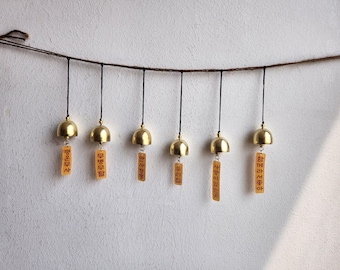 May include: A horizontal string of six gold-colored metal bells hanging from a brown cord. Each bell is attached to a small, rectangular, light-orange paper tag featuring vertical black Korean text characters hanging from the bottom of the bell.