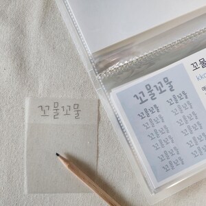 May include: A translucent sticky note with the Korean text 꼬물꼬물 written in black ink, positioned next to a wooden pencil and a clear plastic card holder containing a card with the same text and English definitions of the word.