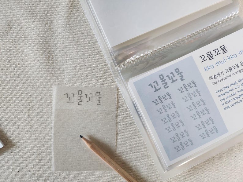 May include: A translucent sticky note with the Korean text 꼬물꼬물 written in black ink, positioned next to a wooden pencil and a clear plastic card holder containing a card with the same text and English definitions of the word.