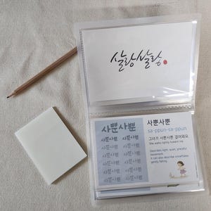 May include: A clear plastic card holder containing white cards with Korean text and illustrations, a wooden pencil, and a stack of translucent sticky notes. The cards feature the text "살랑살랑" and "사뿐사뿐" with English definitions and a small drawing of a person.