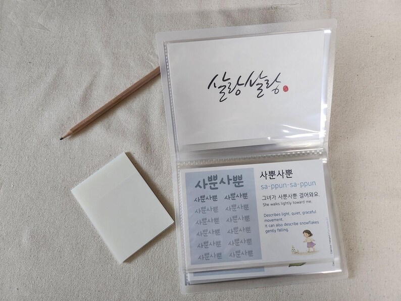 May include: A clear plastic card holder containing white cards with Korean text and illustrations, a wooden pencil, and a stack of translucent sticky notes. The cards feature the text "살랑살랑" and "사뿐사뿐" with English definitions and a small drawing of a person.