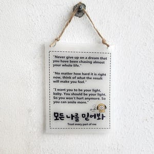 May include: A clear, rectangular sign with a rope hanger, featuring inspirational text in English and Korean. The sign reads "Never give up on a dream..." and "Trust every part of me."