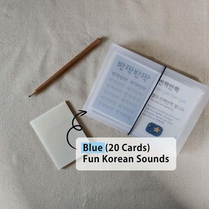 May include: A set of 20 Blue Fun Korean Sounds cards in a translucent frosted plastic case secured with a black elastic band, accompanied by a small matching translucent sleeve and a wooden pencil. The cards feature the Korean text 반짝반짝.