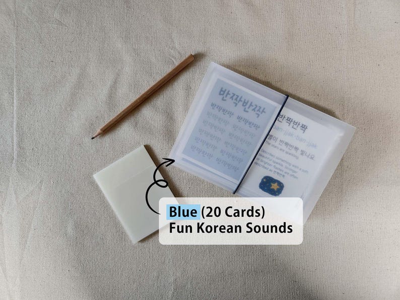 May include: A set of 20 Blue Fun Korean Sounds cards in a translucent frosted plastic case secured with a black elastic band, accompanied by a small matching translucent sleeve and a wooden pencil. The cards feature the Korean text 반짝반짝.