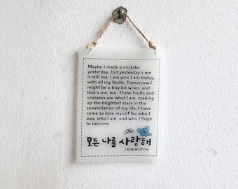 May include: A rectangular white hanging sign with a dashed border, featuring black printed text that reads: Maybe I made a mistake yesterday, but yesterday's me is still me. I am who I am today, with all my faults. Tomorrow I might be a tiny bit wiser, and that's me, too. These faults and mistakes are what I am, making up the brightest stars in the constellation of my life. I have come to love myself for who I was, who I am, and who I hope to become. Below the text is a small blue cartoon character and Korean text.