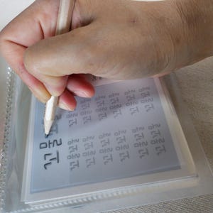 May include: A light-colored wooden pencil held to trace black Korean Hangul characters printed on a white practice sheet. The sheet is encased in a clear plastic protective sleeve, showing multiple rows of repetitive text for writing practice.