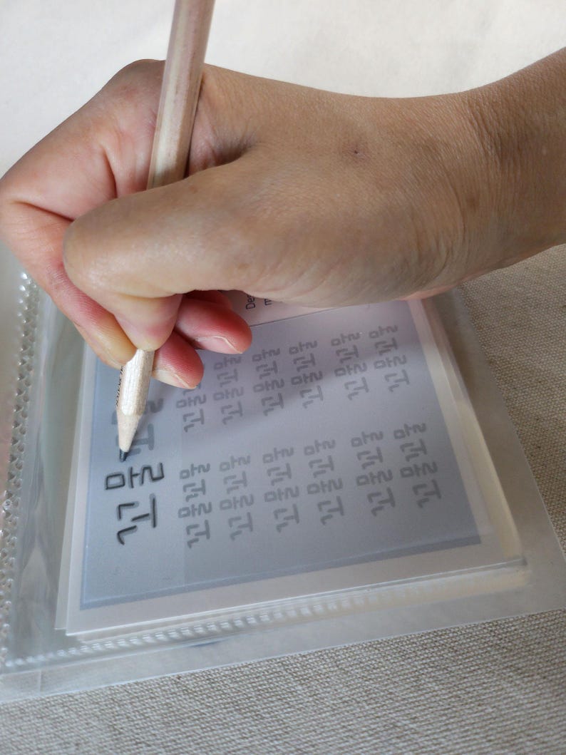 May include: A light-colored wooden pencil held to trace black Korean Hangul characters printed on a white practice sheet. The sheet is encased in a clear plastic protective sleeve, showing multiple rows of repetitive text for writing practice.