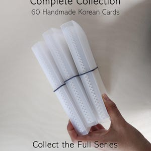 May include: Three translucent white plastic cases containing cards, held together by a thin black elastic band. The text reads Complete Collection 60 Handmade Korean Cards and Collect the Full Series. The cases have a visible white spine detail.