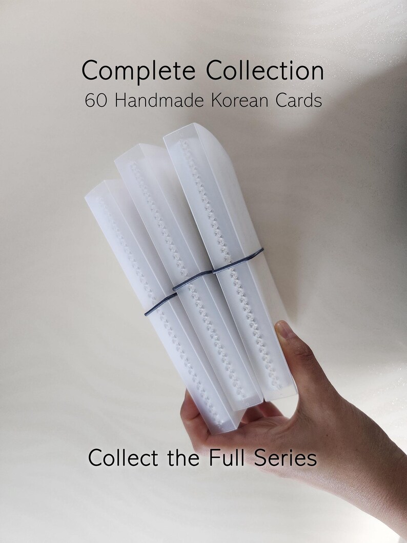 May include: Three translucent white plastic cases containing cards, held together by a thin black elastic band. The text reads Complete Collection 60 Handmade Korean Cards and Collect the Full Series. The cases have a visible white spine detail.
