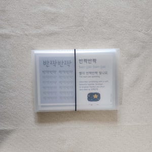 May include: A rectangular, translucent plastic case secured with a thin black elastic band, containing a stack of cards. The visible card features the Korean text '반짝반짝', the romanization 'ban-jjak-ban-jjak', and an illustration of a yellow star.