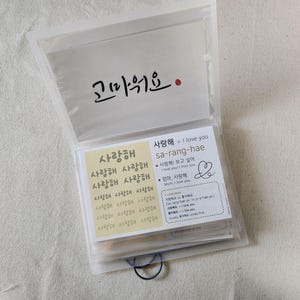 May include: A translucent plastic folder containing multiple cards with Korean text. The top card features the word '고마워요' in black calligraphy. Below, a card displays '사랑해' repeatedly in a grid, alongside a card explaining the phrase 'sa-rang-hae' with English translations and a hand-drawn heart symbol.