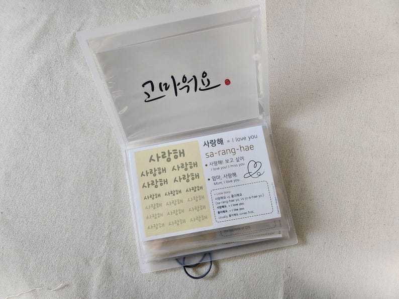 May include: A translucent plastic folder containing multiple cards with Korean text. The top card features the word '고마워요' in black calligraphy. Below, a card displays '사랑해' repeatedly in a grid, alongside a card explaining the phrase 'sa-rang-hae' with English translations and a hand-drawn heart symbol.