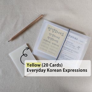 May include: A set of 20 yellow Everyday Korean Expressions cards housed in a translucent plastic case with a black elastic band closure, accompanied by a wooden pencil and a small white card featuring a black arrow graphic pointing to the set.