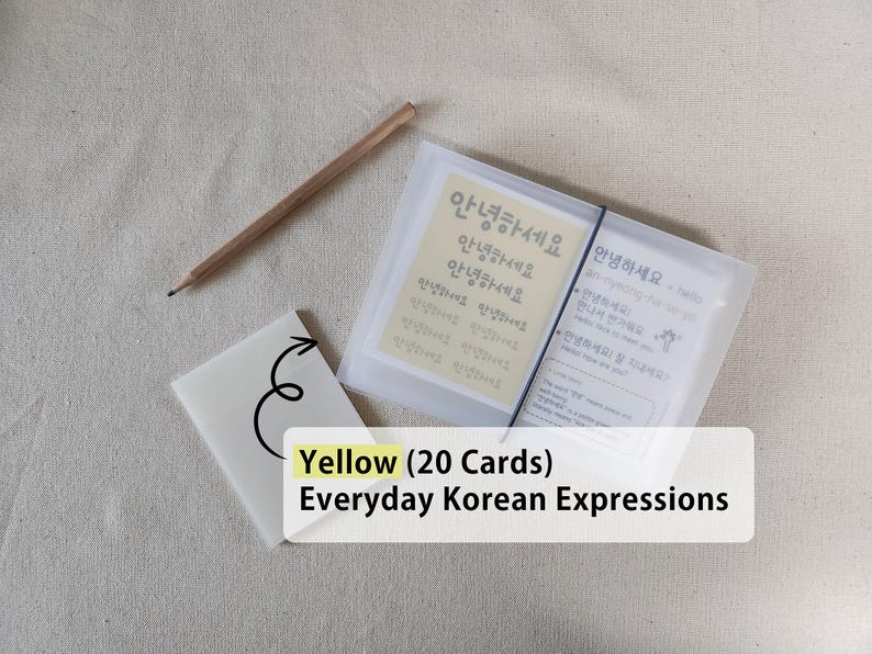 May include: A set of 20 yellow Everyday Korean Expressions cards housed in a translucent plastic case with a black elastic band closure, accompanied by a wooden pencil and a small white card featuring a black arrow graphic pointing to the set.