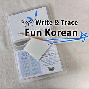 May include: A clear plastic binder containing educational cards for learning Korean, featuring the text Write & Trace Fun Korean. A wooden pencil rests on top of a white rectangular eraser placed over the cards, which display Korean characters.
