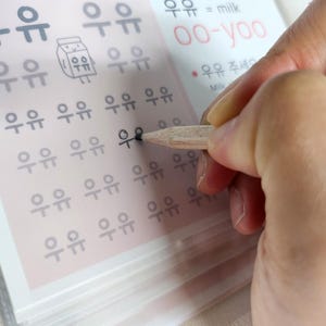 May include: A language learning worksheet featuring rows of the Korean characters for milk, written as 우유, with a small cartoon milk carton illustration, the phonetic pronunciation oo-yoo, and the English translation milk printed in black and red ink.