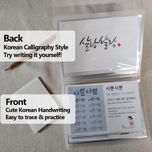 May include: A clear plastic card holder containing instructional cards for learning Korean calligraphy. The cards feature the text '살랑살랑' and '사뿐사뿐' with definitions and practice grids. A wooden pencil and a stack of translucent sticky notes are included.
