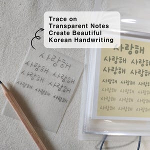May include: A wooden pencil resting on a square piece of transparent paper featuring the Korean text "사랑해" repeated in rows. Next to it is a clear plastic sleeve containing a yellow sheet with printed Korean characters and English definitions.