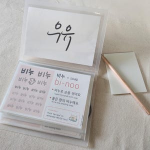 May include: A clear plastic binder containing educational flashcards with Korean text and English translations. One card features the word '비누' repeated with a small soap icon, while another explains '비누 = soap' with example sentences. A wooden pencil lies beside the binder.