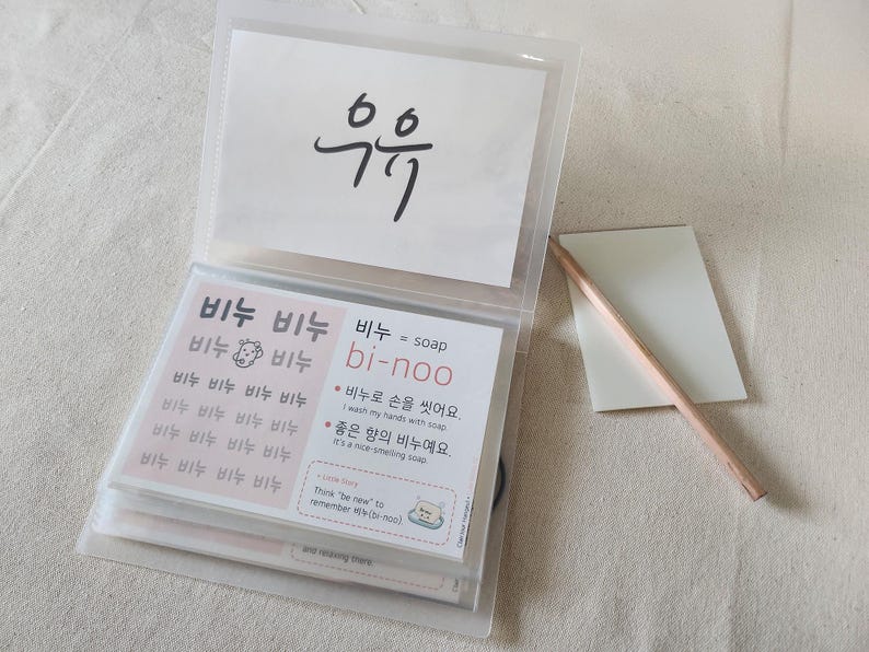 May include: A clear plastic binder containing educational flashcards with Korean text and English translations. One card features the word '비누' repeated with a small soap icon, while another explains '비누 = soap' with example sentences. A wooden pencil lies beside the binder.