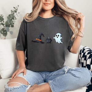 May include: Dark gray t-shirt featuring Halloween-themed graphics. The design includes a witch's hat, a bubbling cauldron, and a friendly ghost. The shirt is made of a soft material and has a relaxed fit, perfect for casual wear during the spooky season.