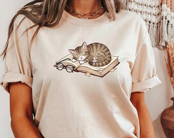 May include: Beige t-shirt featuring a graphic of a sleeping tabby cat curled up on an open book with eyeglasses. The book has a bookmark and the cat is brown and gray. The shirt has short sleeves.