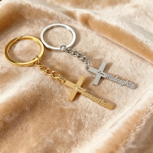 Pode incluir: Dois chaveiros em forma de cruz. Um é dourado com o nome "Samantha" gravado. O outro é prateado com o nome "Midnight" gravado. Ambos os chaveiros têm um anel para chaves.