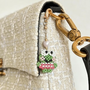 Puede incluir: Un charm de rana de cuentas con una falda rosa y blanca, cuerpo verde y cabeza blanca, colgando de una cadena dorada. El charm está sujeto a un bolso crema con herrajes dorados.