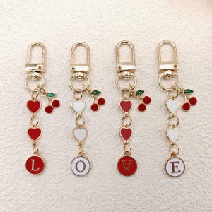 Puede incluir: Cuatro llaveros dorados con dijes de corazón rojos y blancos y detalles de cerezas. Cada llavero tiene una letra diferente: L, O, V y E, en un círculo rojo o blanco.