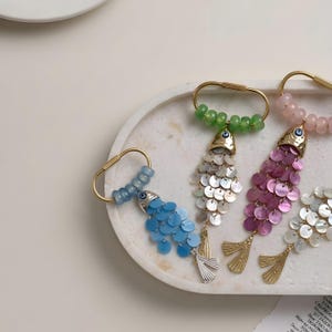 以下が含まれることがあります： 青い邪視のアクセントが付いたゴールドトーンの金属製の頭部を持つ、4つの魚の形をしたキーホルダー。各キーホルダーは、青、緑、ピンク、または白の層状の円形ディスクで構成された本体を持ち、一致するビーズが付いたゴールドトーンの金属リングに取り付けられています。
