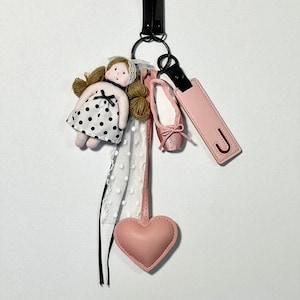 Op de afbeelding: Een sleutelhanger met een pop in een polkadotjurk, een roze balletschoen, een roze hart en een leren label met de letter "J". De sleutelhanger heeft een zwarte leren band en een metalen ring.