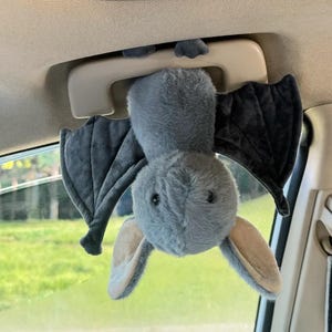 May include: A plush gray bat toy with black wings and tan ears hangs from a car's interior handle. The bat is made of soft material and has a friendly expression. The toy is designed to hang in a vehicle.