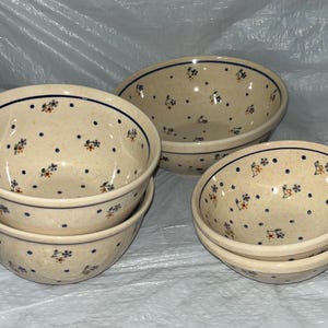 Pode incluir: Um conjunto de tigelas de cerâmica com base creme, adornadas com um padrão repetitivo de pontos azuis e motivos florais em azul, laranja e verde. As tigelas têm uma borda azul escura.