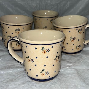 Puede incluir: Cuatro tazas de cerámica con asas. Las tazas tienen una base color crema con lunares azules y pequeños diseños florales en azul, amarillo y naranja. Los bordes y las asas están ribeteados con una línea azul oscuro.