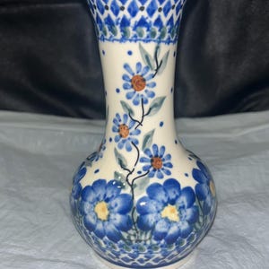 Puede incluir: Un jarrón de cerámica con base color crema que presenta flores azules pintadas a mano con centros amarillos y naranjas. El cuello y la base están decorados con un patrón de diamantes azul y blanco, con pequeños puntos azules dispersos.