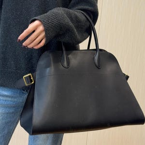 Può includere: Una borsa nera dal design strutturato, con un manico superiore e una tracolla con fibbia color oro. La borsa è tenuta da una persona che indossa un maglione scuro con scollo a V e jeans blu.