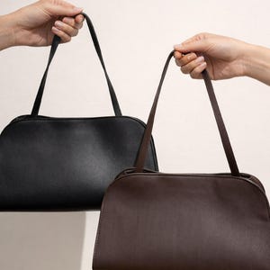 May include: Two shoulder bags are held up against a white background. One bag is black, and the other is brown. Both bags have a rectangular shape with rounded edges and long, thin straps.