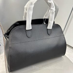 May include: A black leather handbag with a textured finish. The bag has a rounded shape, a top handle wrapped in white paper, and a silver chain detail on the side. The bag is sitting on a gray chair.